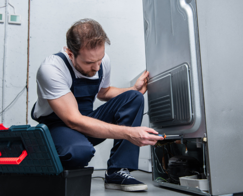 Side view, adult bearded repairman in working overall fixing refrigerator by screwdriver in kitchen