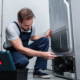 Side view, adult bearded repairman in working overall fixing refrigerator by screwdriver in kitchen